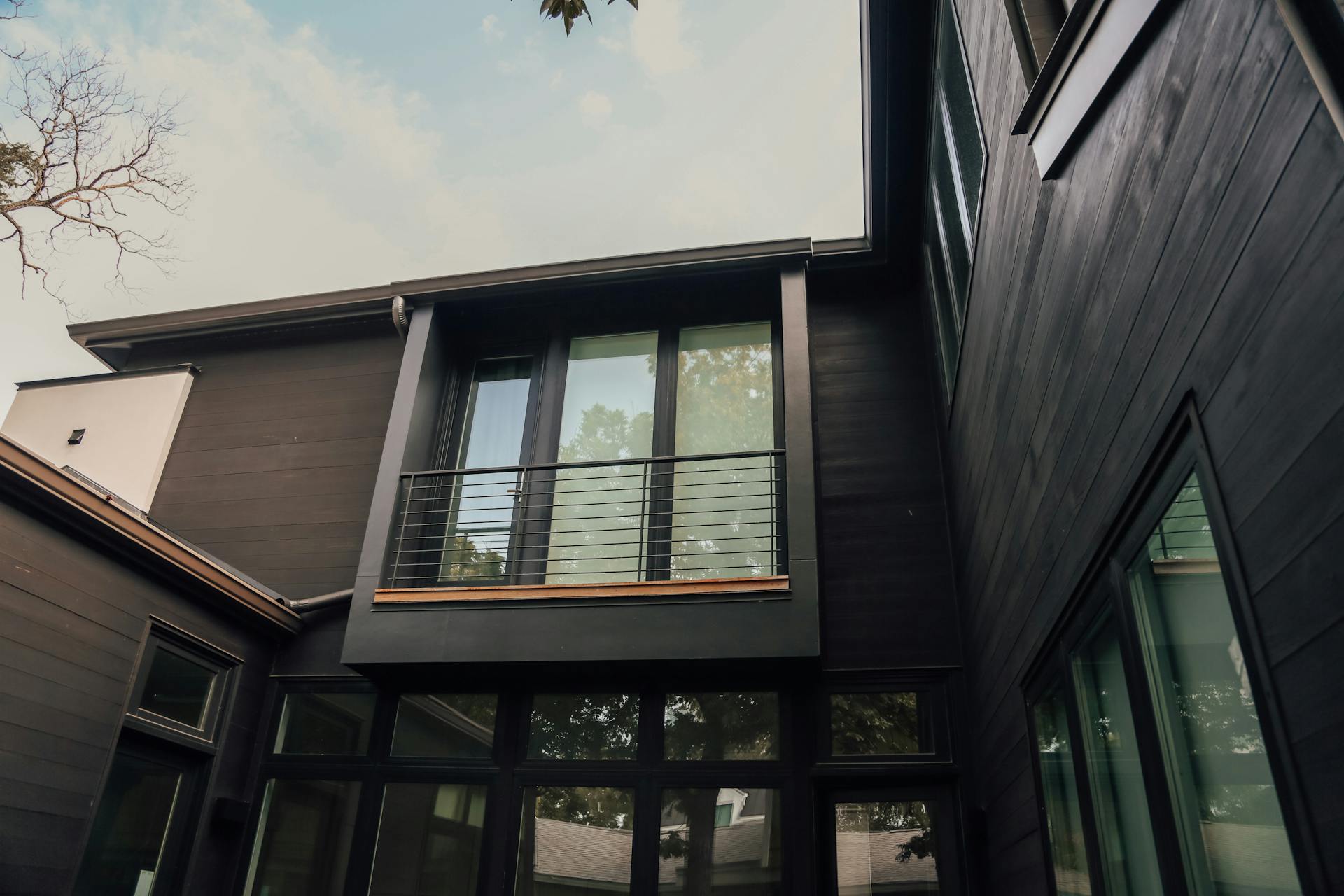 From below of simple open balcony of tall luxury cottage with black walls constructed of wood under cloudy sky and leafless branches