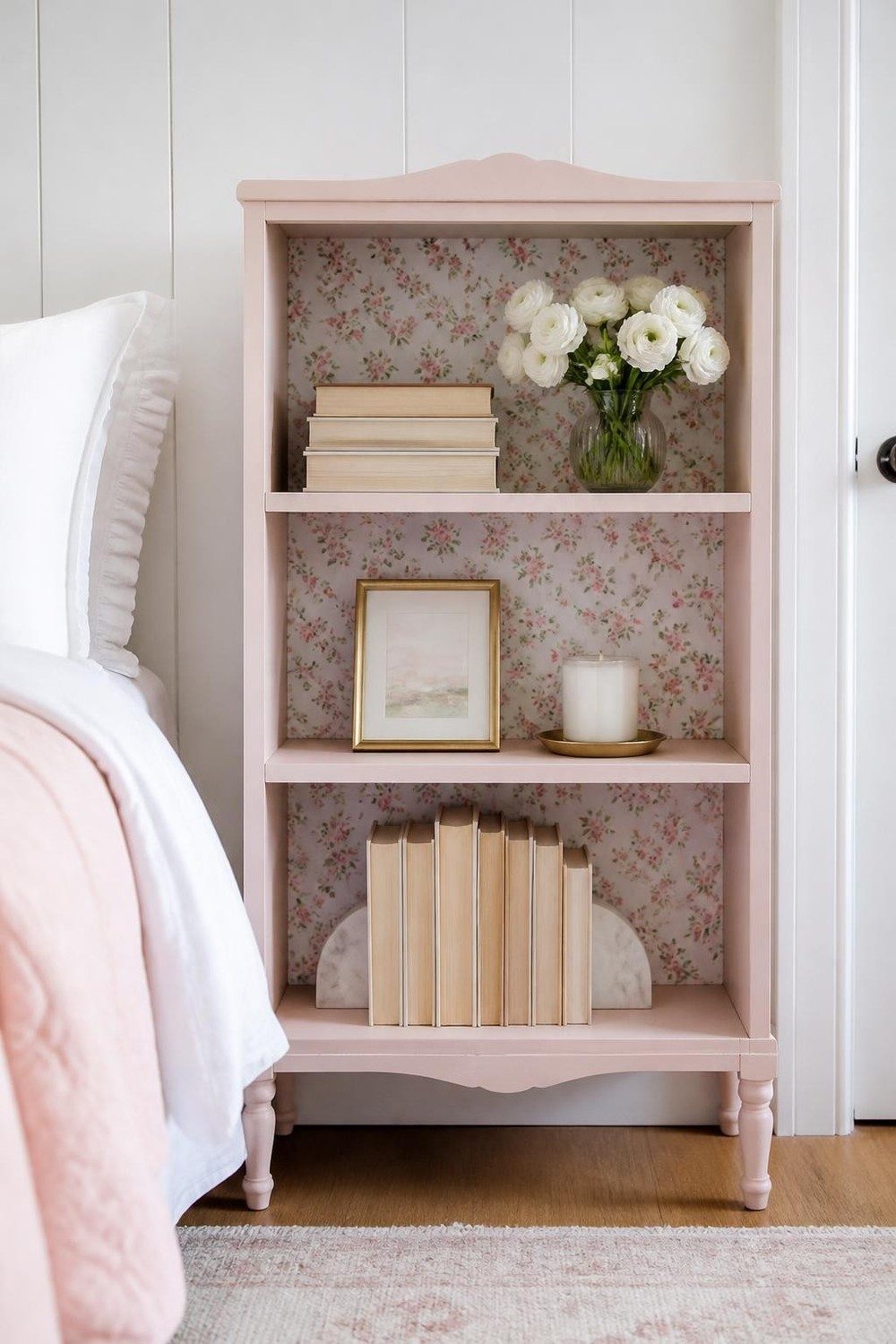 Bedroom bookcase lined with tiny blush floral wallpaper holding novels and white flowers