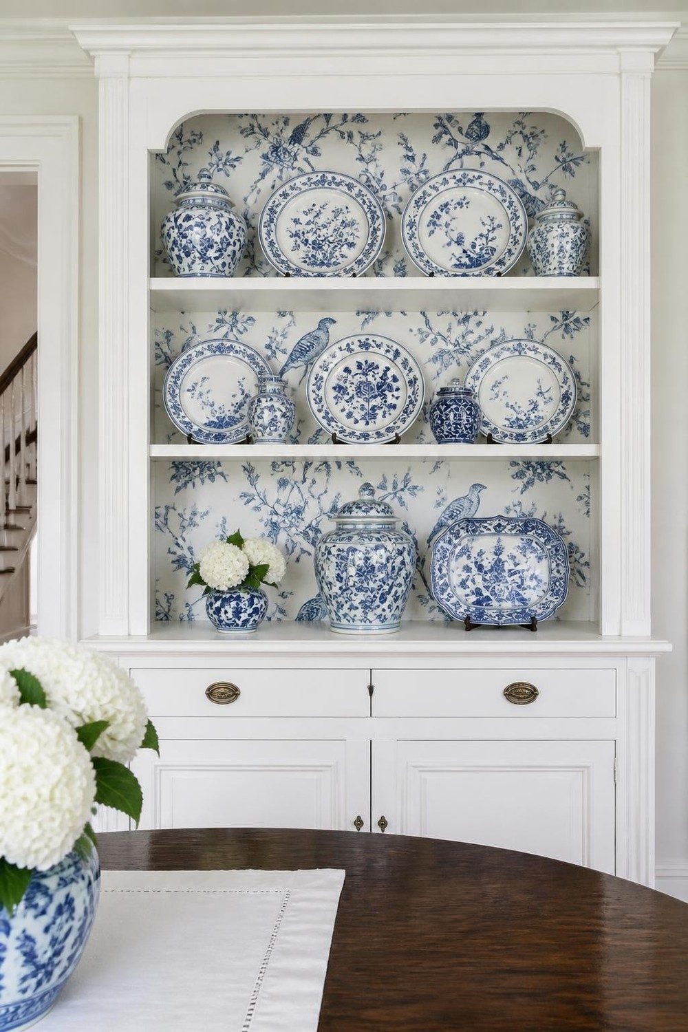 Dining room hutch lined with blue and white chinoiserie wallpaper displaying blue and white china