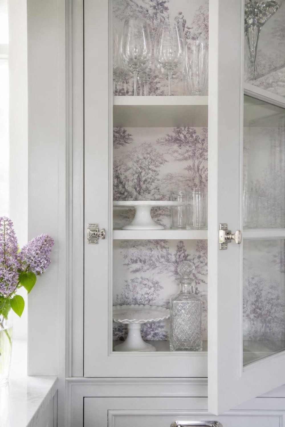 Gray glass-front cabinet lined with lavender toile wallpaper and crystal glassware